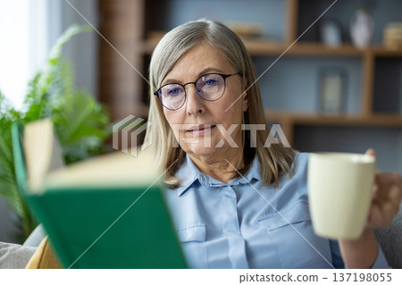 Senior woman wearing glasses reading book while drinking coffee at home. Relaxed elderly lady enjoying quiet time and leisure activity in cozy living room environment. Senior woman wearing glasses reading book while drinking coffee at home. Relaxed elderly lady enjoying quiet time and leisure activity in cozy living room environment. 137198055