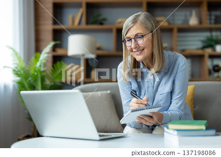 Mature woman studying online with laptop, writing notes, smiling while learning at home. Lifelong learning, education, and remote studying concept 137198086