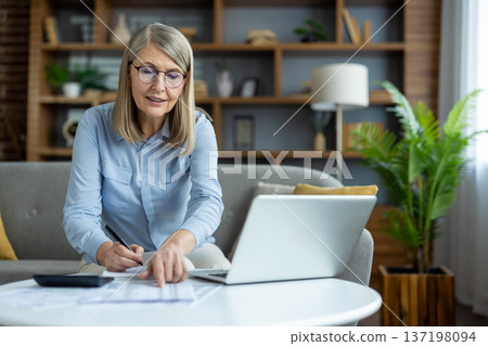 Senior woman working from home on laptop, reviewing documents and using calculator in modern living room. Home office setup with desk and bookshelf in background. Concept of remote work productivity. 137198094