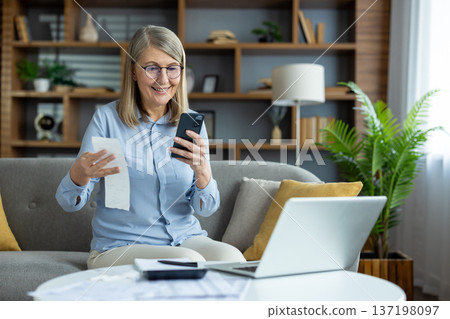Older woman sitting on sofa using smartphone and holding receipt. Managing household budget, paying utilities and rent payments online. Happy and relaxed at home. Older woman sitting on sofa using smartphone and holding receipt. Managing household budget, paying utilities and rent payments online. Happy and relaxed at home. 137198097