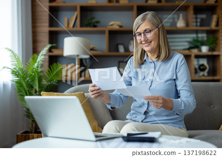 Senior woman sitting on couch reviewing documents while working from home on laptop. Appears focused and content in cozy living room setting, engaged in remote work or personal tasks Senior woman sitting on couch reviewing documents while working from home on laptop. Appears focused and content in cozy living room setting, engaged in remote work or personal tasks 137198622