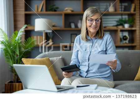 Senior woman sitting on couch, using laptop and reviewing financial documents. Concept of home finances, budget management, and senior independent living. Senior woman sitting on couch, using laptop and reviewing financial documents. Concept of home finances, budget management, and senior independent living. 137198623