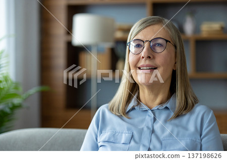 Smiling mature woman with glasses looking happy while sitting in cozy living room. Modern home interior with bookshelf and lamp in background 137198626