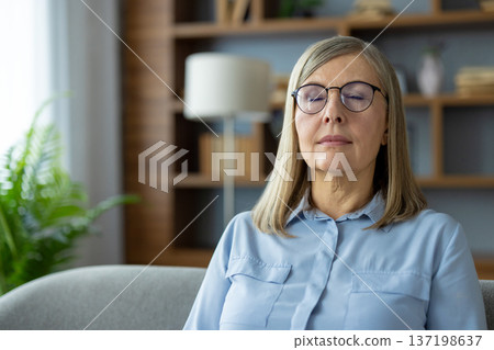Senior woman with glasses sitting on couch practicing mindfulness and meditation. Eyes closed, calm expression. Serenity, peace, and relaxation during self-care at home. Senior woman with glasses sitting on couch practicing mindfulness and meditation. Eyes closed, calm expression. Serenity, peace, and relaxation during self-care at home. 137198637