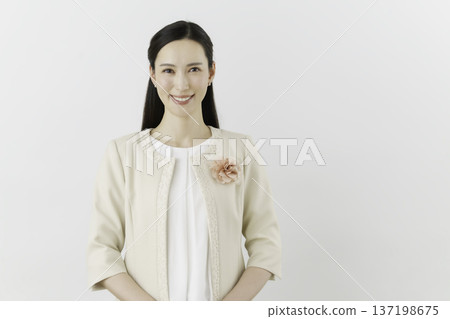 A young Japanese woman wearing a white blouse and cream-colored jacket against a white background, wearing a flower corsage on her chest and smiling 137198675