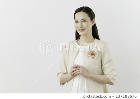 A woman wearing a white blouse and cream jacket against a white background, with a flower corsage on her chest and hands clasped A woman wearing a white blouse and cream jacket against a white background, with a flower corsage on her chest and hands clasped 137198676