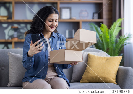 A woman smiles while holding her phone and opening a delivery box, showcasing her excitement. She is sitting on a sofa in a cozy living room environment. 137198826