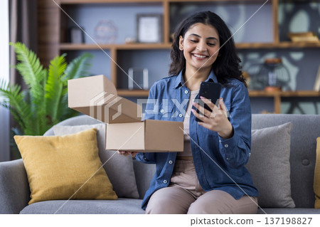 Smiling young Indian woman sitting on the couch at home, holding a received parcel, an open box and looking at the mobile phone screen. Smiling young Indian woman sitting on the couch at home, holding a received parcel, an open box and looking at the mobile phone screen. 137198827