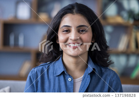 Close-up portrait of a young smiling Indian woman in a denim shirt indoors, standing and smiling at the camera. Close-up portrait of a young smiling Indian woman in a denim shirt indoors, standing and smiling at the camera. 137198828