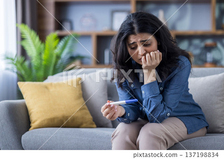 A woman looks anxiously at a pregnancy test, sitting on a couch in a living room. This image conveys uncertainty about the future. A woman looks anxiously at a pregnancy test, sitting on a couch in a living room. This image conveys uncertainty about the future. 137198834