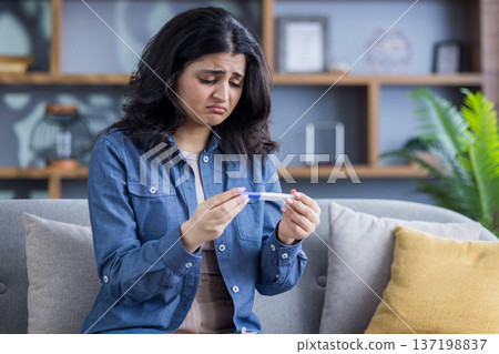 Upset young Muslim woman sitting on the couch at home and looking disappointedly at the rapid pregnancy test she is holding in her hands. 137198837