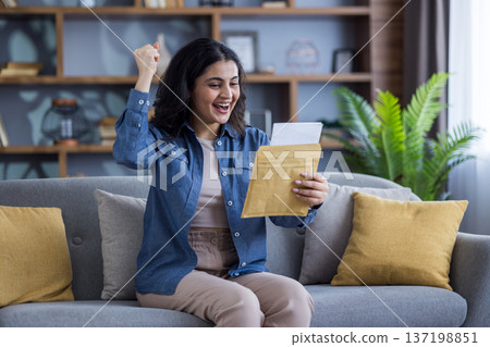 Happy Indian young woman rejoices at the letter she received, sits at home on the couch, raises her hand up, holds an open envelope. 137198851