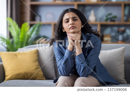 Portrait of a sad and worried young Muslim woman sitting on the couch at home, folding her hands together and looking sadly at the camera. 137199006