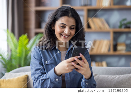 Close-up photo of a smiling young Muslim woman sitting on the couch at home, holding a phone and looking at the screen. 137199295