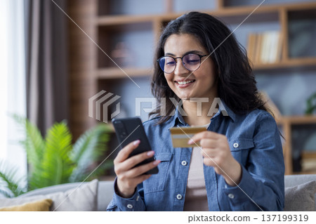 Close-up photo of a young Indian woman sitting on the sofa at home, holding a credit card and using a mobile phone. Close-up photo of a young Indian woman sitting on the sofa at home, holding a credit card and using a mobile phone. 137199319
