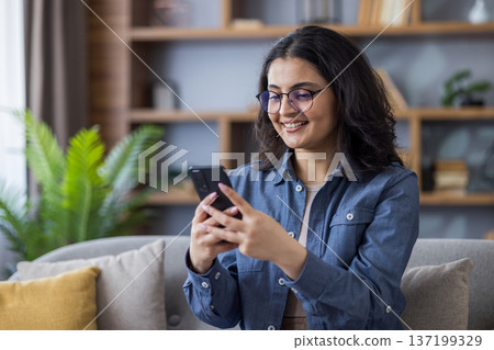 Smiling young Indian woman sitting on sofa at home and using mobile phone. Smiling young Indian woman sitting on sofa at home and using mobile phone. 137199329