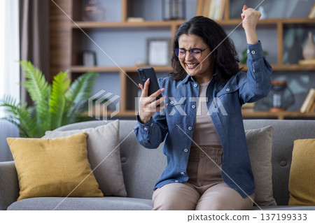 A happy woman on a couch raises her arm in celebration after looking at her phone. She's smiling and wearing glasses. A happy woman on a couch raises her arm in celebration after looking at her phone. She's smiling and wearing glasses. 137199333