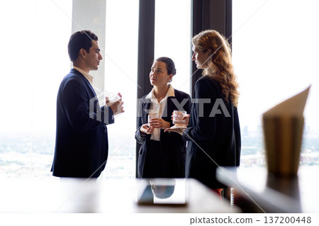 Business professionals networking by a large window at a corporate event, discussing strategy and partnership while holding drinks in a modern office setting with city view background Business professionals networking by a large window at a corporate event, discussing strategy and partnership while holding drinks in a modern office setting with city view background 137200448