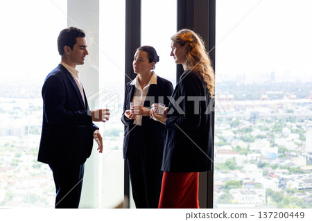 Business professionals discussing strategy during a corporate networking event by a large office window with city view, holding drinks and building partnership in a modern urban workplace Business professionals discussing strategy during a corporate networking event by a large office window with city view, holding drinks and building partnership in a modern urban workplace 137200449