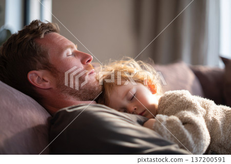 dad leaning back on couch with toddler resting against him, eyes closed, soft warm sunlight, neutral living room interior, vertical composition 137200591