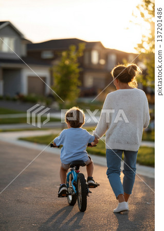 mother teaching her child to ride a bicycle in suburban neighborhood, supportive gesture, soft evening sunlight, modern houses in background, emotional milestone moment, vertical framing 137201486