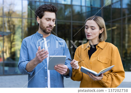 Business professionals collaborating and having a serious discussion outdoors, working on a project with a digital tablet and notebook in front of a modern building Business professionals collaborating and having a serious discussion outdoors, working on a project with a digital tablet and notebook in front of a modern building 137202738