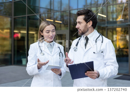 Two doctors confer outside a modern clinic, reviewing a tablet and clipboard as they discuss patient care, diagnosis and treatment plans in a professional teamwork setting Two doctors confer outside a modern clinic, reviewing a tablet and clipboard as they discuss patient care, diagnosis and treatment plans in a professional teamwork setting 137202740