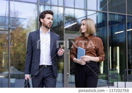 Two professional business colleagues, a man and a woman, are walking and engaging in conversation outside a modern corporate office building, discussing work Two professional business colleagues, a man and a woman, are walking and engaging in conversation outside a modern corporate office building, discussing work 137202768