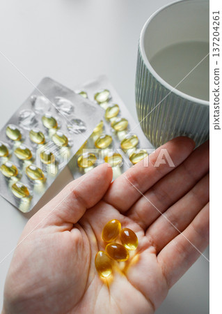 Omega 3 fish oil capsules in woman hand and in blisters on white table with cup of water. Vitamins and supplements, healthcare and medicine. 137204261