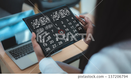 Over the shoulder view of a business person using a digital tablet with a calendar app to schedule appointments and manage deadlines at a workplace with a laptop on a wooden desk. 137206794