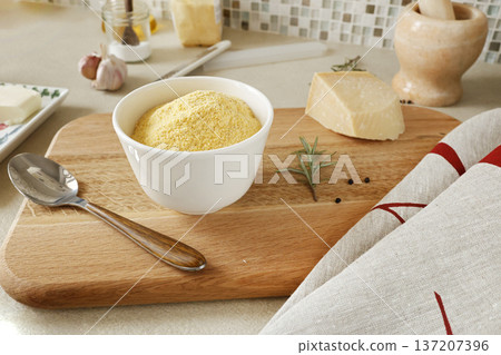 Corn flour in white ceramic bowl on wooden cutting board in kitchen. Parmesan and rosemary. Ingredients for polenta. Traditional Italian cuisine. 137207396