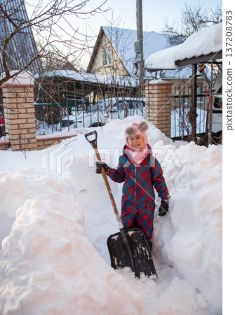 Young girl shoveling snow with large shovel in snowy yard near trees and fence in winter countryside 137208783