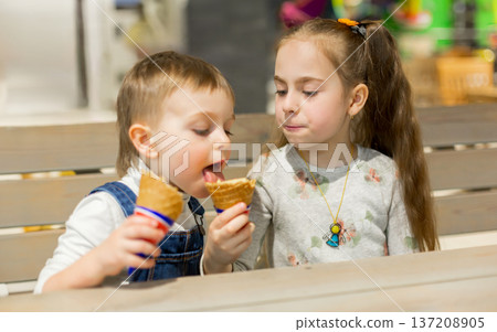 Two children sharing ice cream cone while sitting at wooden table indoors in family entertainment center Two children sharing ice cream cone while sitting at wooden table indoors in family entertainment center 137208905