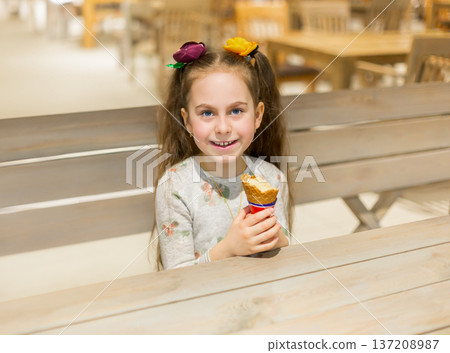 Smiling little girl holding ice cream cone while sitting wooden table cozy cafe with warm lighting 137208987