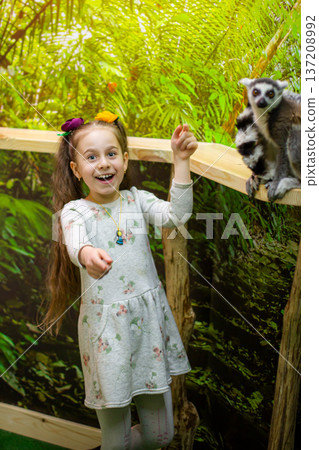 Generated imageHappy little girl pointing ring tailed lemur wooden railing in tropical indoor zoo with background 137208992
