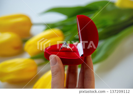 Hand holding open red ring box with diamond ring against blurred yellow tulips background romantic Hand holding open red ring box with diamond ring against blurred yellow tulips background romantic 137209112