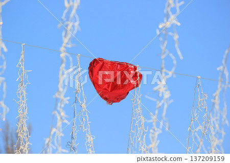 Red heart foil balloon among white garland lights against a clear blue sky background. 137209159