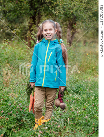 Smiling girl standing orchard holding carrots beets, wearing jacket during autumn harvest outdoors 137209382