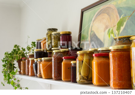 Canned vegetables and fruits are lined up on a shelf in a kitchen, showing winter preparations. A painting of a basket is seen in the background as part of the decor 137210407