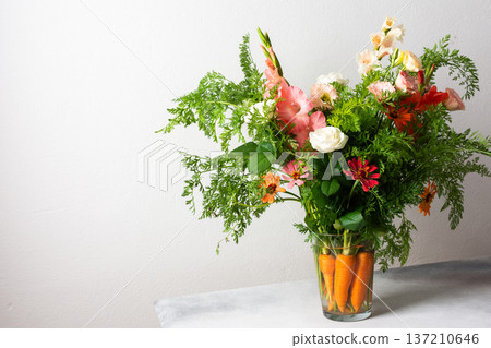 A bouquet of roses, gladioli, zinnias, and eustomas stands in water. Carrots with green tops are part of the arrangement on a plain table against a light background 137210646