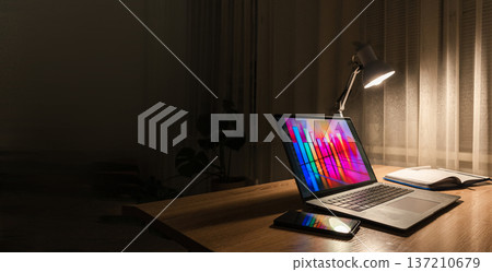 A desk lamp illuminates a laptop on a wooden table at night, displaying a colorful histogram and data graph 137210679