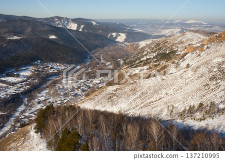 Aerial view of a village nestled in a snowy valley with mountain peaks. Winter landscape with forest and houses for holiday vacation background. Torgashinsky ridge, Krasnoyarsk, Siberia 137210995