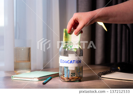 Saving for education fund using money jar. Young person carefully placing money into labeled Education savings jar. Perfect for student finance, parental planning, and investment in knowledge themes 137212533