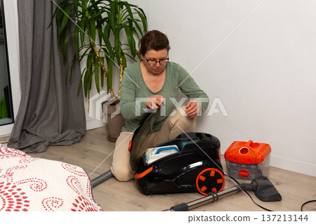 In a living room, a person is getting a vacuum cleaner ready for cleaning. The person is seated on the floor with the vacuum cleaner open and looks focused on the task In a living room, a person is getting a vacuum cleaner ready for cleaning. The person is seated on the floor with the vacuum cleaner open and looks focused on the task 137213144