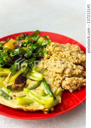 Grains, cooked greens, and vegetables are arranged on a red plate. A bowl of mixed salad with tomatoes is placed nearby on a light surface 137213145