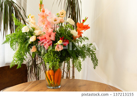 A bouquet features roses, gladioli, zinnias, and eustomas. Carrots with green tops sit in water, placed on a wooden table in a bright room A bouquet features roses, gladioli, zinnias, and eustomas. Carrots with green tops sit in water, placed on a wooden table in a bright room 137213155