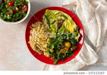 Grains, cooked greens, and vegetables are arranged on a red plate. A bowl of mixed salad with tomatoes is placed nearby on a light surface Grains, cooked greens, and vegetables are arranged on a red plate. A bowl of mixed salad with tomatoes is placed nearby on a light surface 137213167