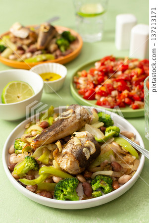 Breakfast includes baked hake placed on a bed of boiled beans with broccoli and peppers, accompanied by water and tomato salad on the table Breakfast includes baked hake placed on a bed of boiled beans with broccoli and peppers, accompanied by water and tomato salad on the table 137213171