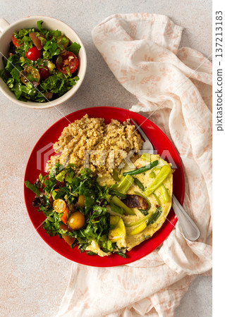 Grains, cooked greens, and vegetables are arranged on a red plate. A bowl of mixed salad with tomatoes is placed nearby on a light surface 137213183