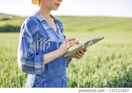 A woman in overalls and a straw hat uses a tablet computer while standing in a green agricultural field 137213307